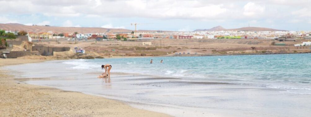 Tranquil coastal city beach with golden sand, gentle waves, cloudy sky, and distant seaside buildings.