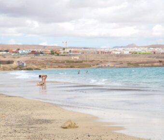 Tranquil coastal city beach with golden sand, gentle waves, cloudy sky, and distant seaside buildings.
