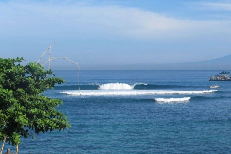 Calm blue ocean swell with breaking waves near tropical pier, distant mountain and lush coastal foliage.
