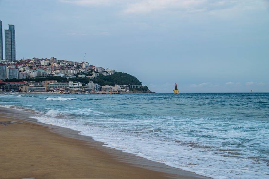 Tranquil sandy beach with gentle waves, coastal city skyline, and yellow navigation buoy in turquoise water.