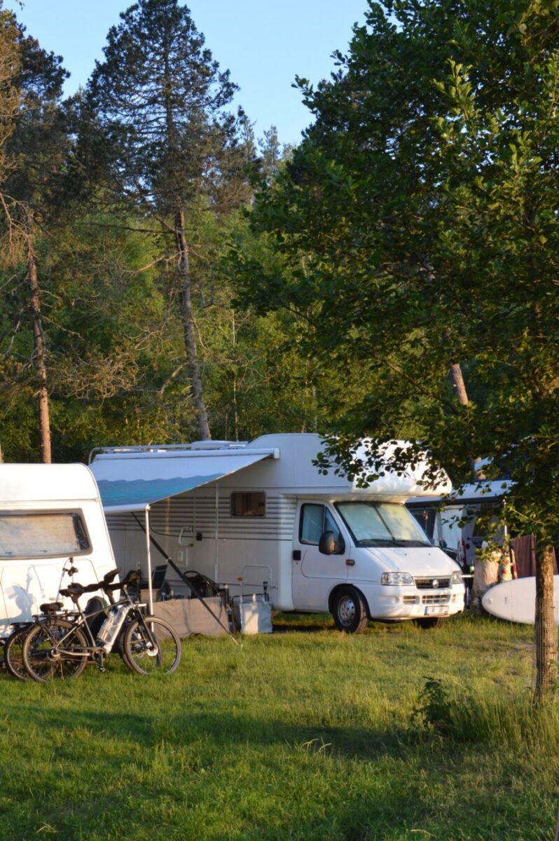 Serene campsite with a white camper van and bicycles under a green awning. Sunlight filters through trees, creating a peaceful and relaxing ambiance.