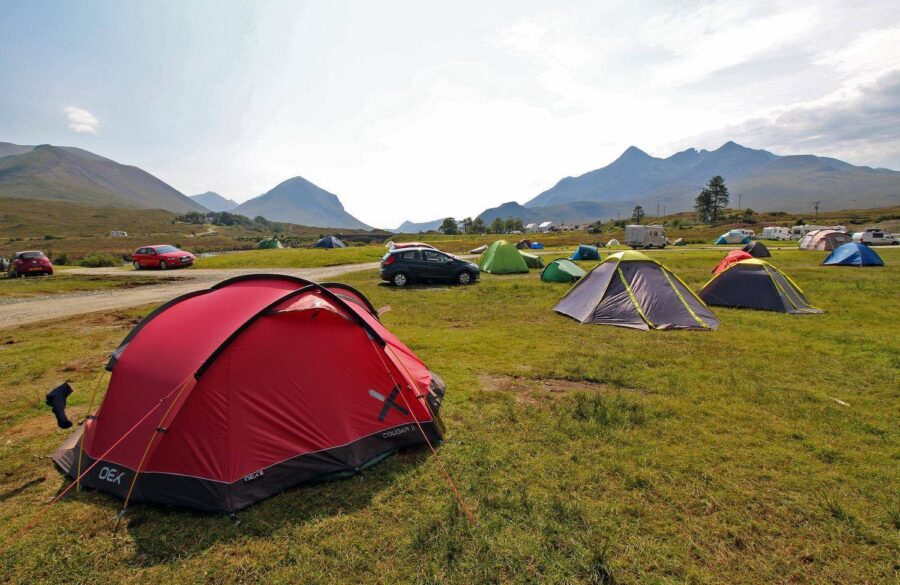 Scenic campsite with colorful tents, majestic mountains, and lush landscape under a clear blue sky.
