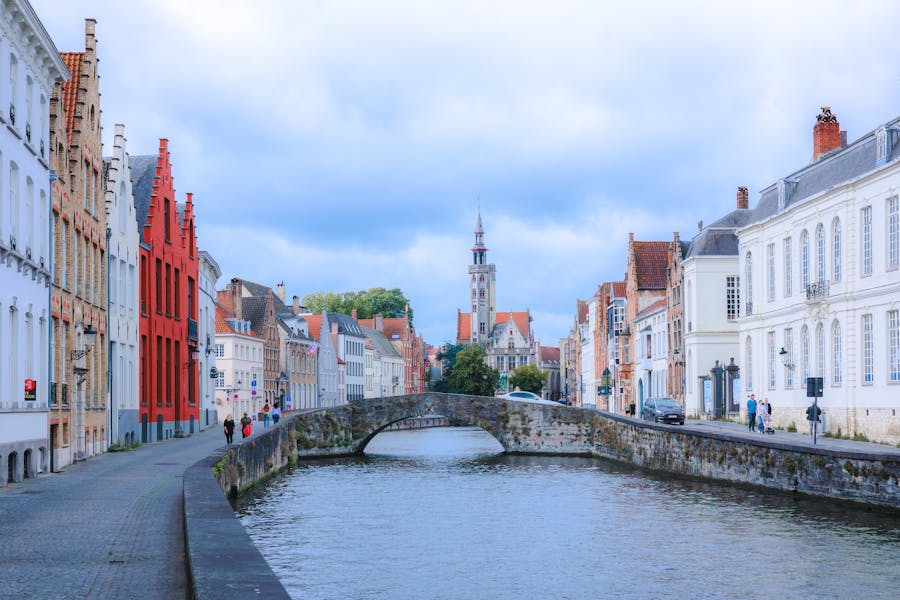 Bruges canal with stone bridge, historic waterfront buildings and bell tower reflected in water.