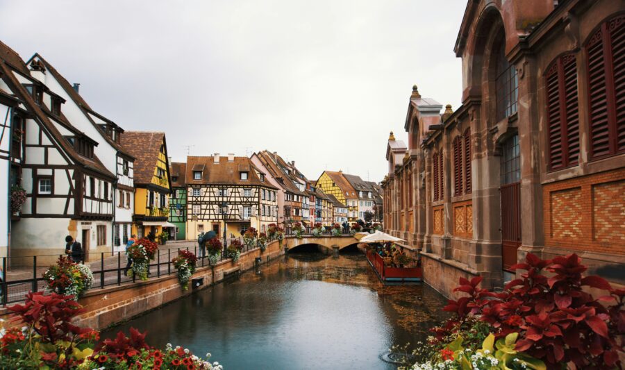 Alsace canal with stone bridge, half-timbered houses and riverside flowers.