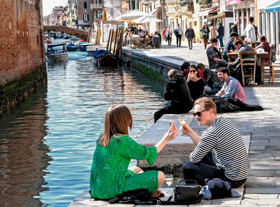 Couple toasting white wine by a sunny canal with boats and waterfront cafes.