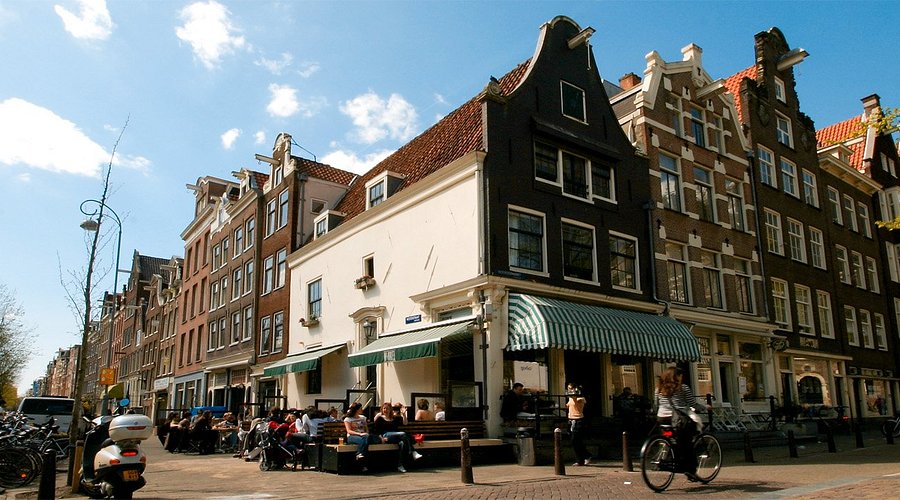 Amsterdam canal-side cafe on cobbled street with historic Dutch gabled houses, cyclists, and pedestrians.
