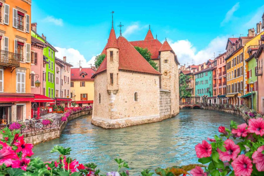 Annecy canal with stone island house, pastel waterfront buildings, and pink flowers.