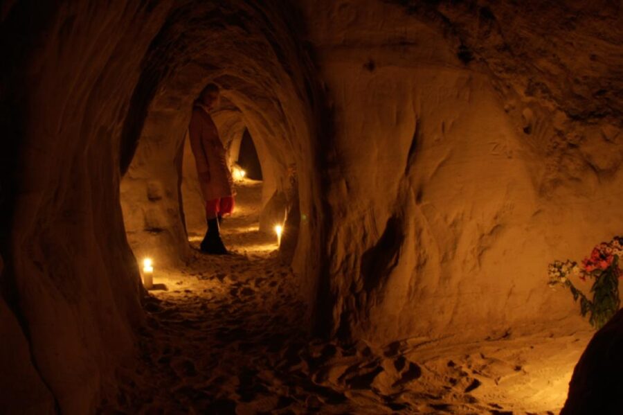 Candlelit cave with figure in long coat, flowers, and sandy walls, evoking introspection.