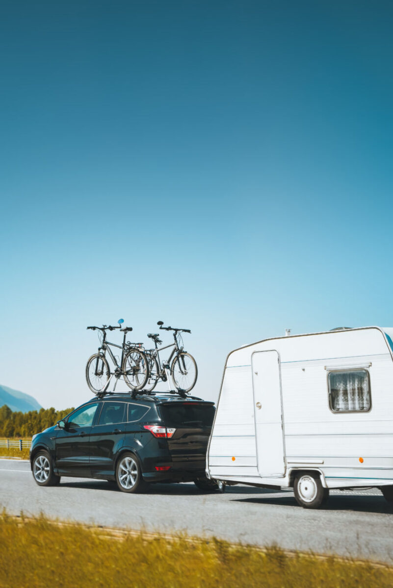 Black car towing a white camper trailer drives on a road under a clear blue sky. Two bicycles are mounted on the car roof, evoking a sense of adventure.