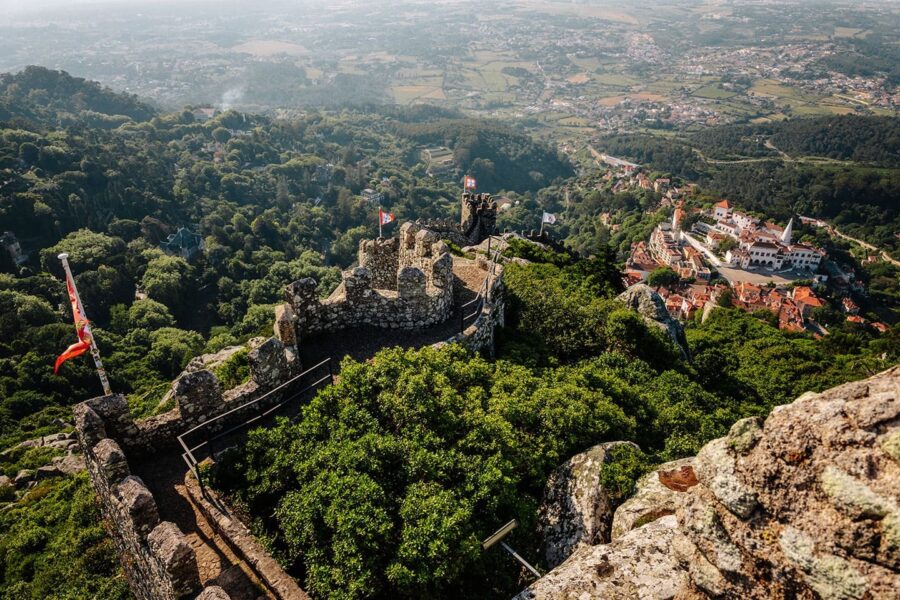 Ancient castle ruins on hilltop with distant town, lush landscape, and fluttering flags.