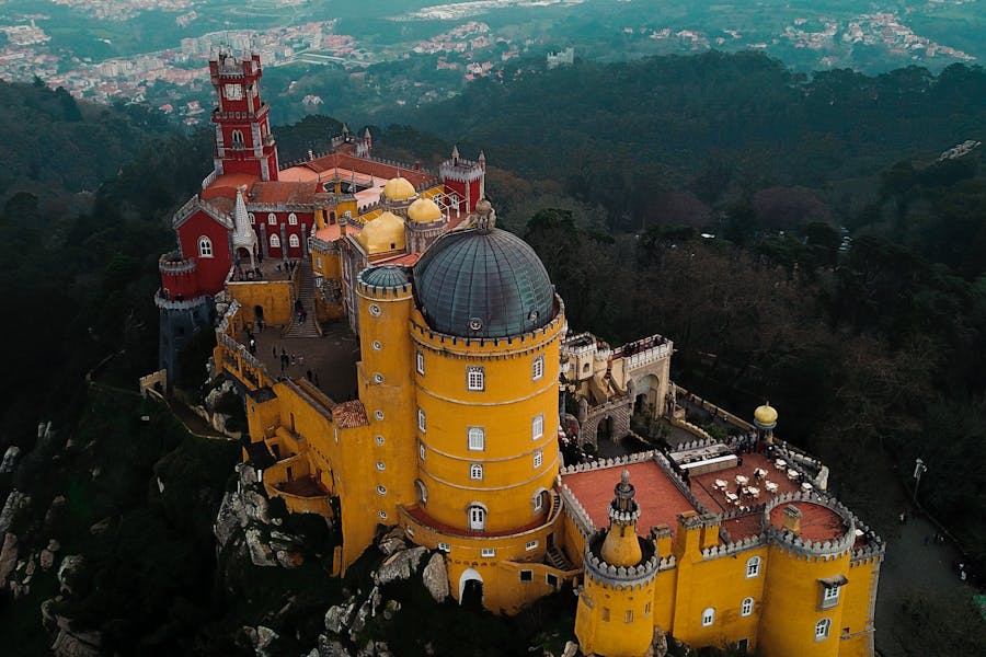 Aerial view of vibrant yellow castle amid lush green hills and distant mountains.