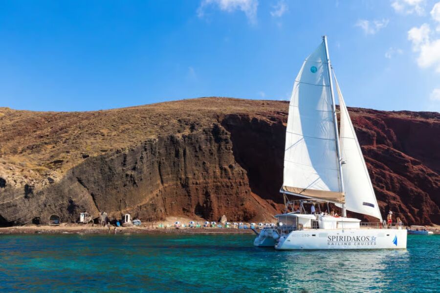 Catamaran glides through the ocean near a cliff, part of a sailing cruise experience from Santorini.
