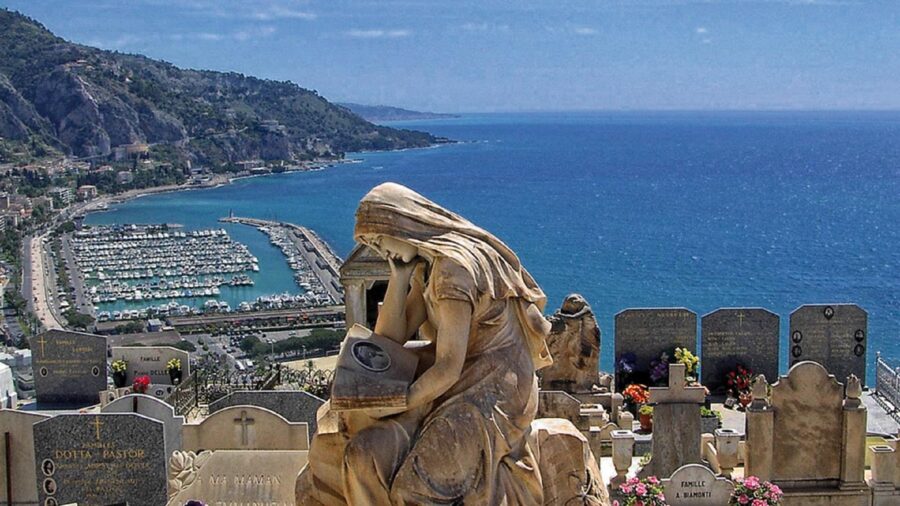 Hillside cemetery with mourning statue overlooking marina and deep blue coastal sea.