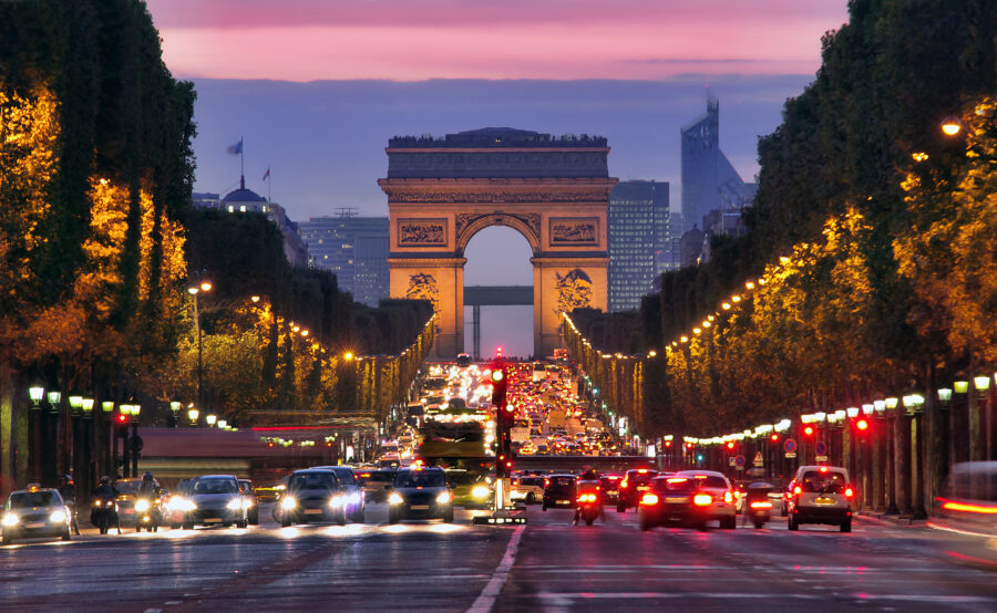Night view of Champs Elysees and Arc de Triomphe in Paris, bustling with car traffic and illuminated city lights.