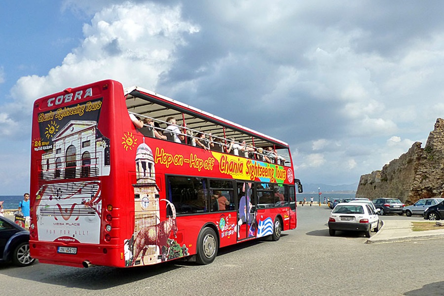 Red double-decker bus tours Chanias stunning coastal landmarks under a sunny sky.