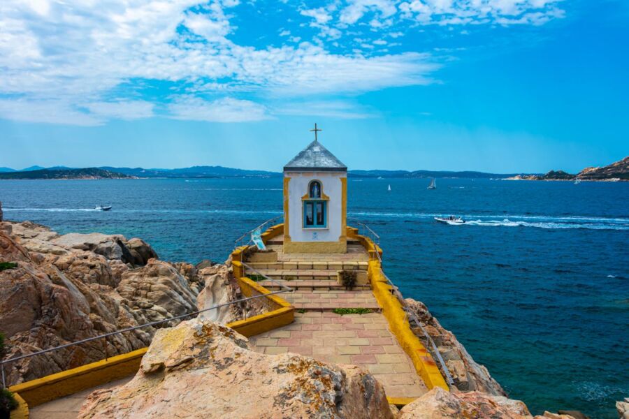 Coastal chapel with yellow facade, rocky outcrop, blue arched windows, ocean, and sailboats.