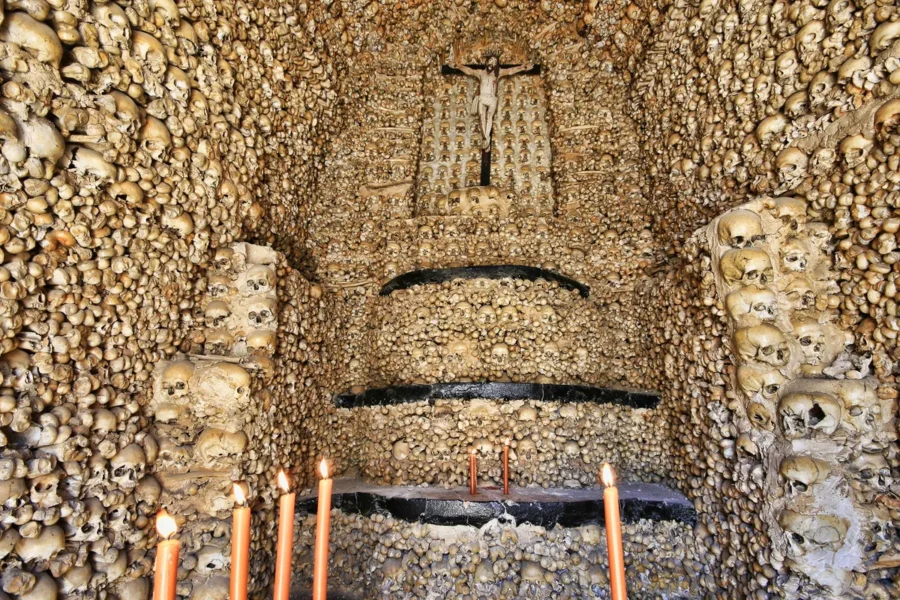 Haunting chapel interior with skulls, bones, crucifix, altar, and candles symbolizing mortality and reflection.