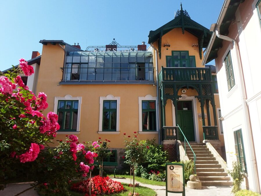 Charming yellow building with green balcony, glass structure, pink roses, and ornate details.
