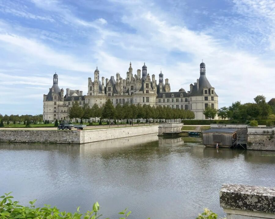 Majestic castle with numerous spires and towers reflected in a calm moat. The sky is bright and partly cloudy, adding a serene atmosphere.