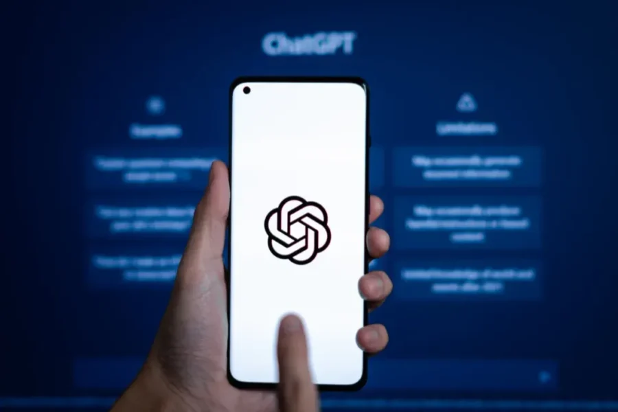 Person using ChatGPT OpenAI mobile app on smartphone in front of computer screen.