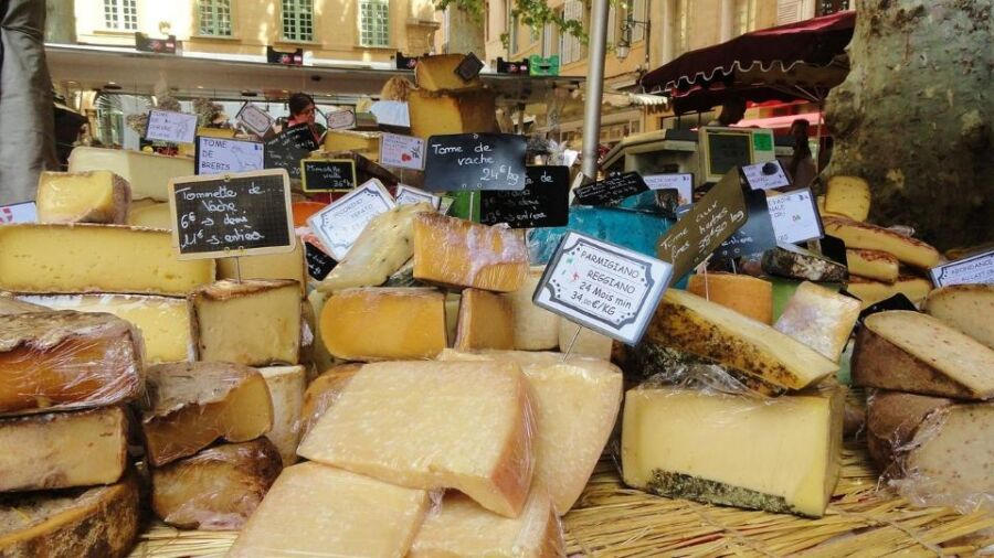 Cheese market stall with diverse artisan cheeses, prices, and bustling shoppers in a quaint setting.