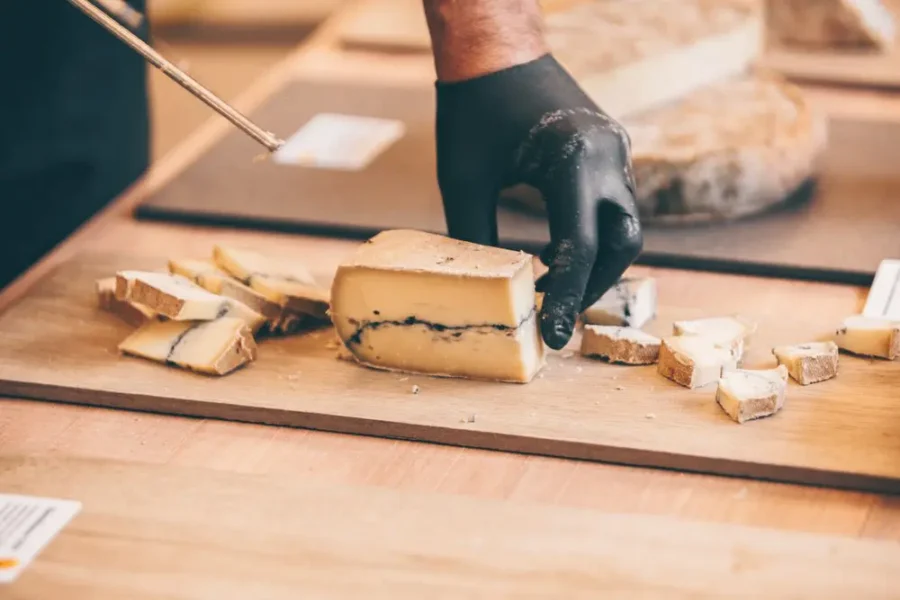 Person slicing gourmet cheese block with dark veins on wooden countertop.
