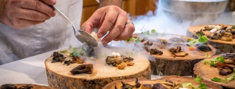 Chef plating gourmet dish with mushrooms, herbs, smoke, on rustic wooden board in kitchen.