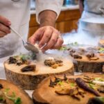 Chef plating gourmet dish with mushrooms, herbs, smoke, on rustic wooden board in kitchen.