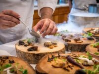 Chef plating gourmet dish with mushrooms, herbs, smoke, on rustic wooden board in kitchen.