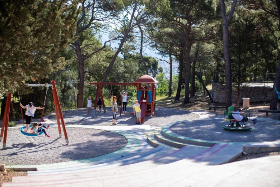 Vibrant playground under pine trees, kids playing, swings, and merry-go-round.