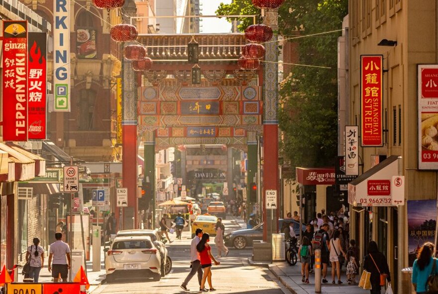 Bustling Chinatown street with ornate Chinese archway, red lanterns, restaurants, signs and pedestrians.