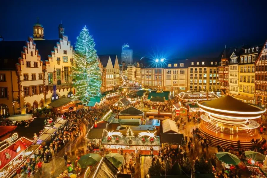 Christmas market in town square at night with lights, crowded stalls, carousel, and tree.