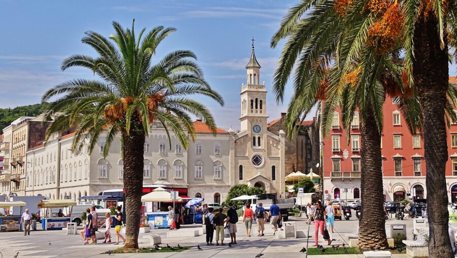Mediterranean square with palm trees, historic church, market stalls, and diverse visitors.