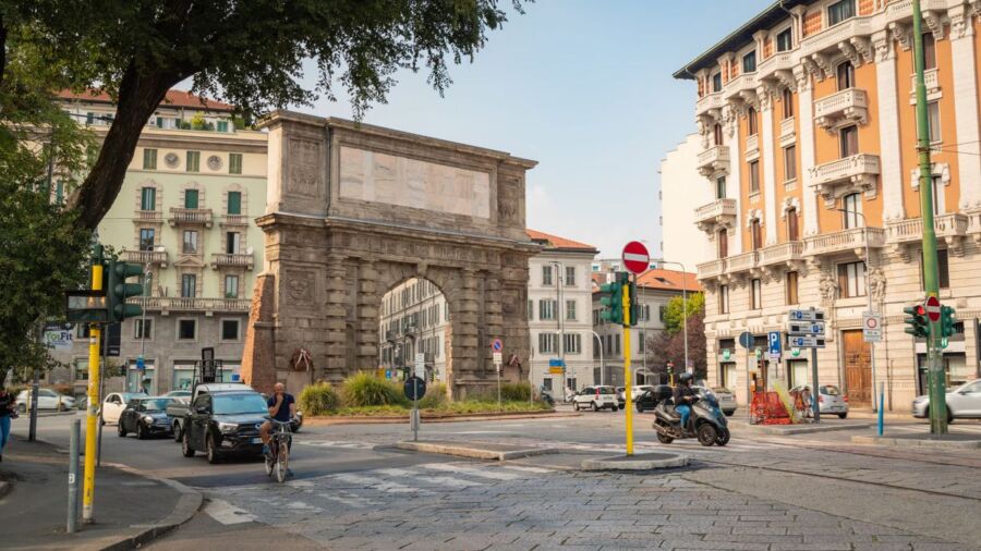 Urban intersection with historical arch, cars, cyclist, and diverse architectural styles in vibrant city.
