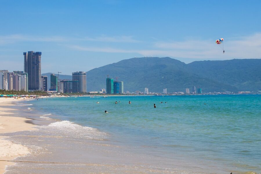 Modern My Khe Beach city skyline with turquoise water, mountains, swimmers, and colorful parasailing.