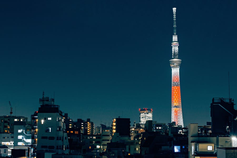 Illuminated broadcast tower rising above dense night city skyline with glowing windows.