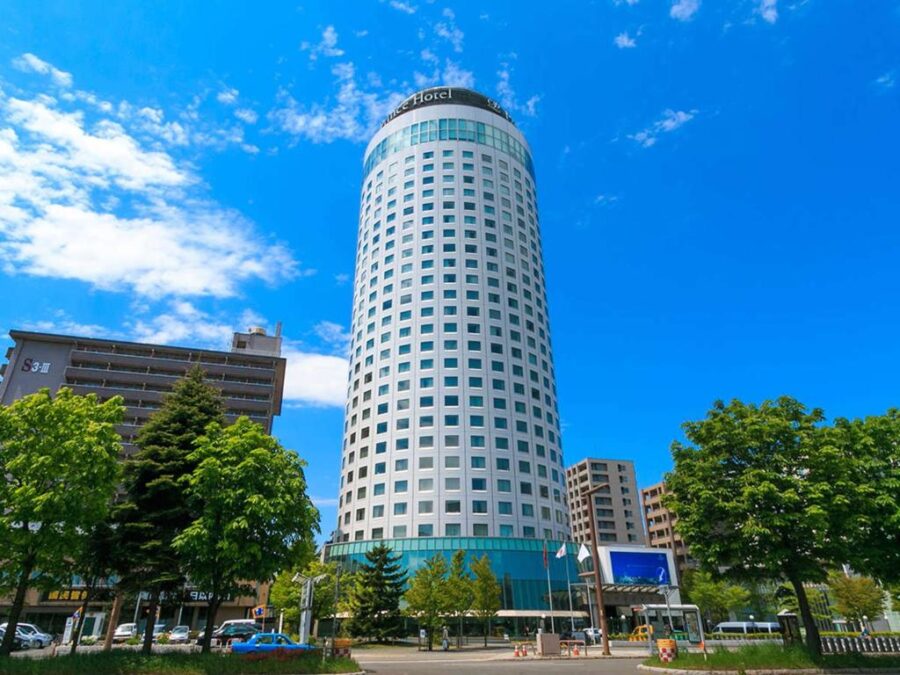 Sapporo Prince Hotel cylindrical tower with windows, flags, trees and traffic under blue sky.