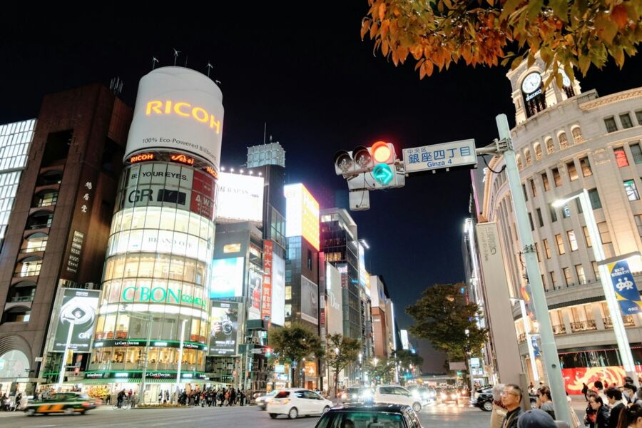 Tokyo Ginza nightlife: vibrant neon lights, bustling crosswalk, Ricoh building, autumn foliage.