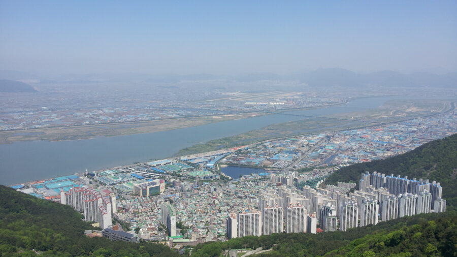 Aerial view of Sasang city skyscrapers, river, industrial district, farmland, and distant mountains.