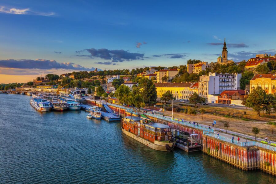 Serene riverside scene in Belgrade with vibrant sunset, boats, and diverse architecture.
