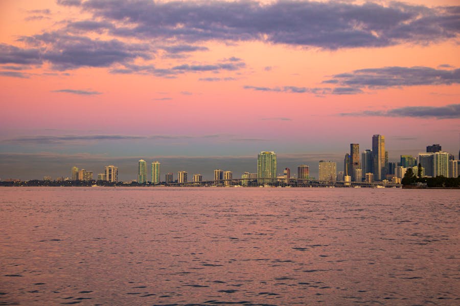 City skyline at sunset over calm waterfront with pastel reflections.