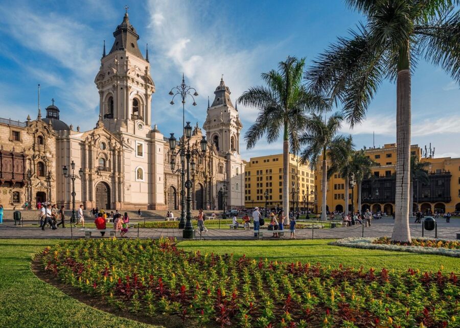 Historic city square with colonial architecture, floral gardens, and palm trees under a blue sky.
