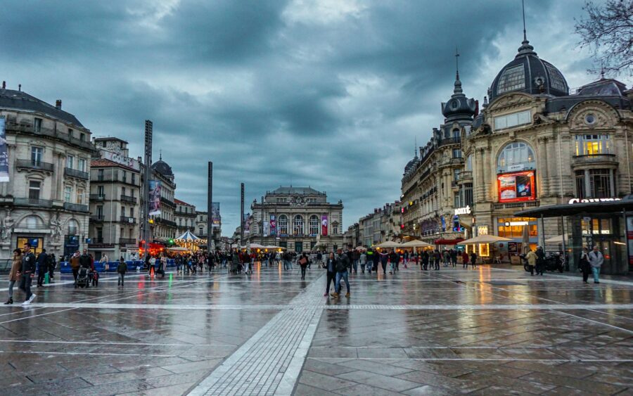 Montpellier city square at rainy dusk with wet paving, historic buildings, carousel and pedestrians.