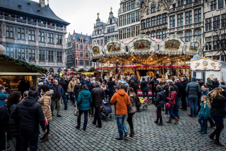 Winter city square carousel with festive lights, historic buildings, and bustling market crowd.