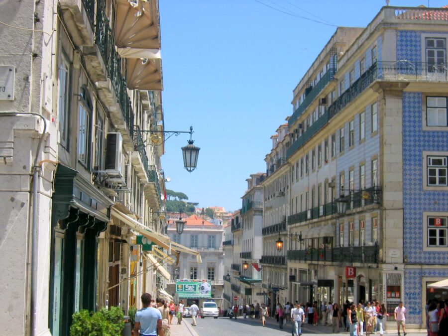 Sunlit Rua Garrett city street in Lisbon with cafes, tiled facades, balconies, pedestrians, and terracotta rooftops.