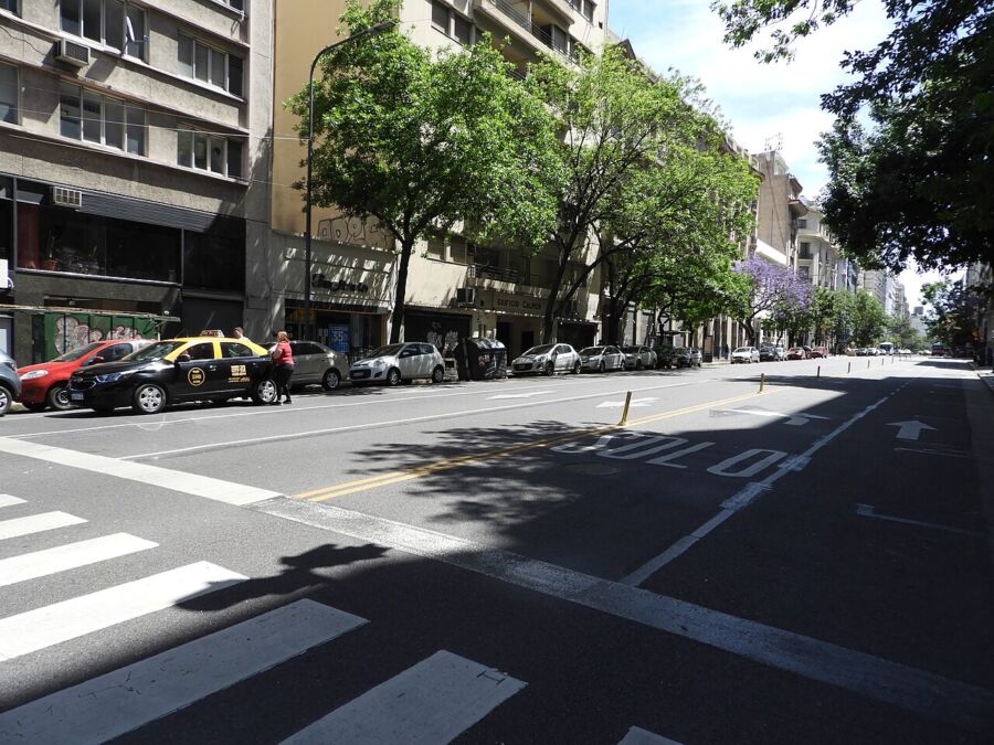 Belgrano street with yellow taxi, parked cars, mixed architecture, graffiti, and greenery.