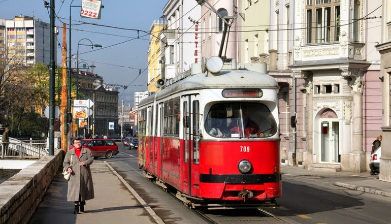 Red vintage Sarajevo tram 709 on sunny European city street with pedestrians and historic buildings.