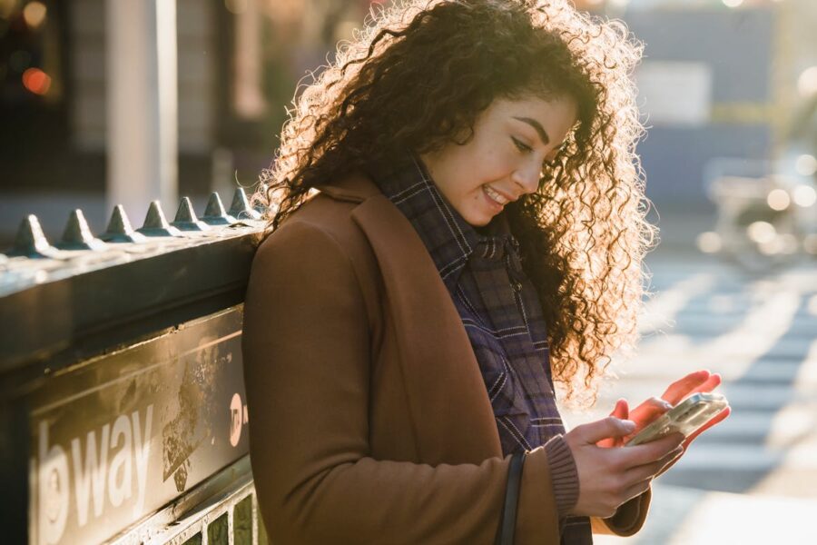 Young woman smiling, checking smartphone in sunlit urban scene near subway.