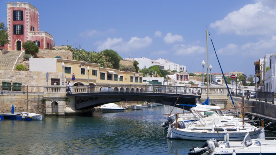 Picturesque bridge crosses a river in Ciutadella, Menorca, surrounded by lush greenery and clear blue skies.