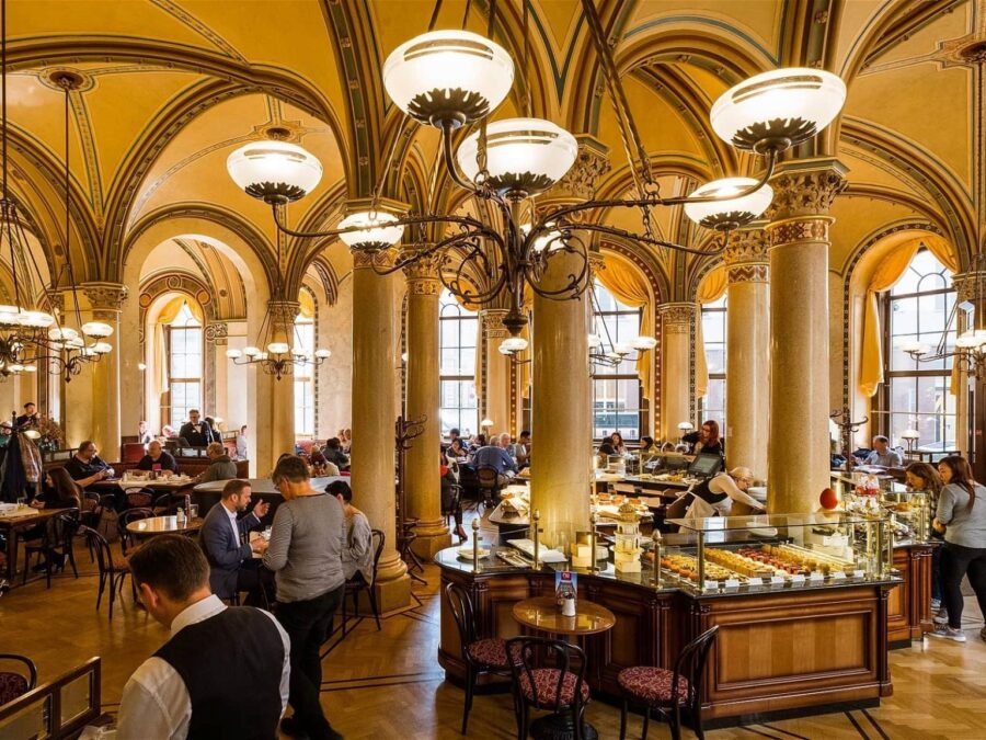 Historic café interior with ornate columns, chandeliers, pastry display, and patrons.
